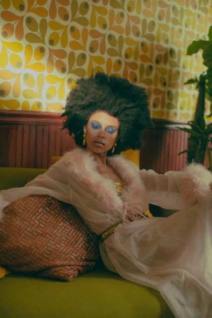 Woman with afro and blue eye makeup lounges in sheer robe on green couch with patterned pillow in front of patterned yellow wallpaper.
