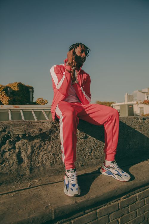A young adult male in a red tracksuit sits on a ledge outdoors on a sunny day, posing with his hands near his face.