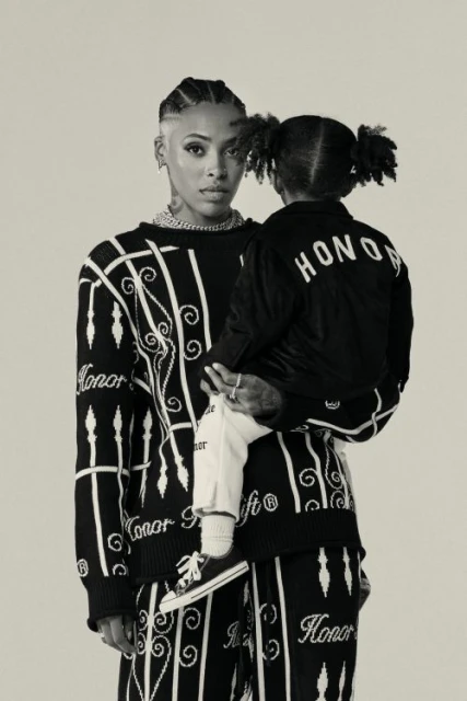 Adult and child fashion portrait. Woman in patterned knitwear holds toddler with pigtails wearing matching black "Honor" jacket.
