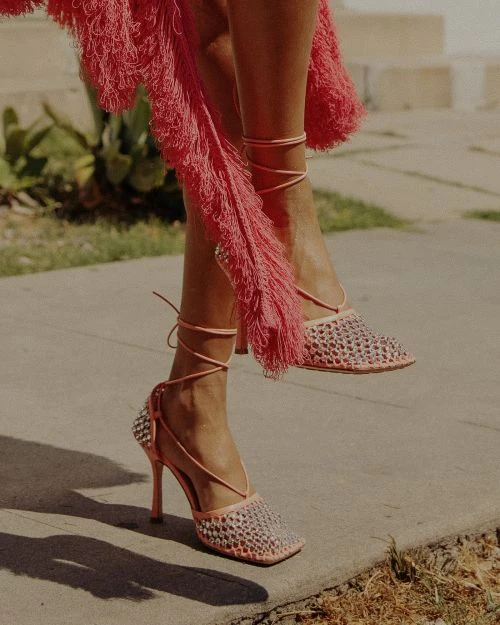 Close-up of fashion-forward woven heels with pink fringe detail, photographed outdoors on a concrete surface with leafy greens in the background.
