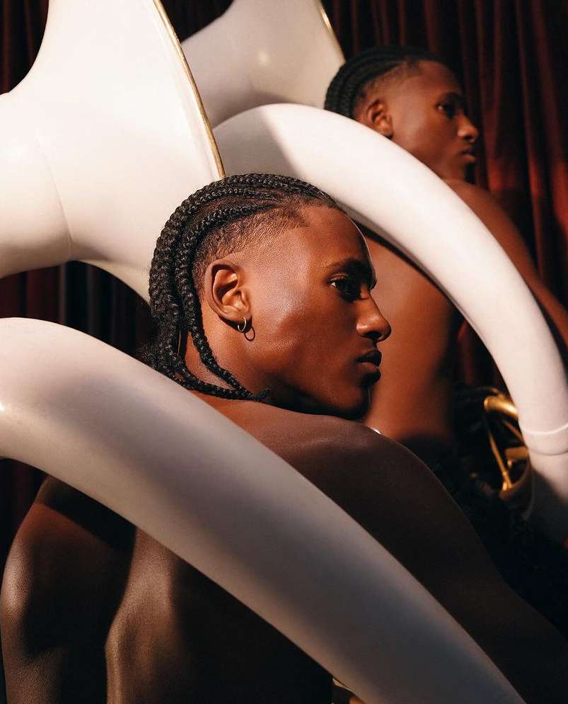 Two Black men pose shirtless with white inflatable swan objects in a studio setting with dramatic lighting and a red curtain backdrop.