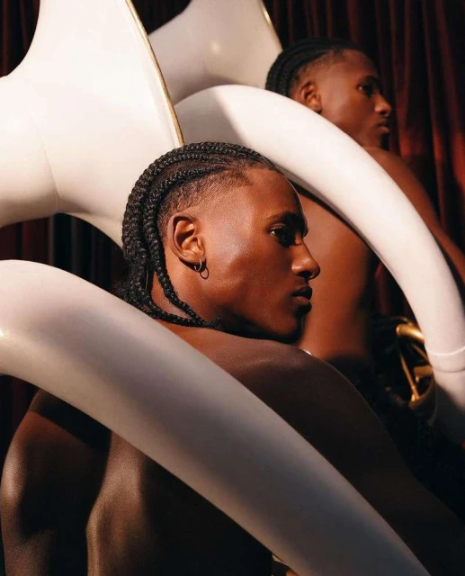 Two Black men pose shirtless with white inflatable swan objects in a studio setting with dramatic lighting and a red curtain backdrop.