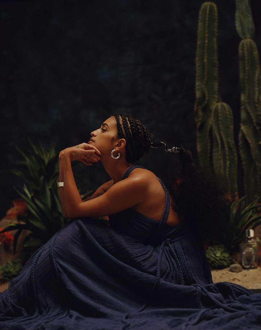 Elegant woman in flowing blue dress poses thoughtfully in a desert-like setting with cacti and dark background, illuminated by soft light.