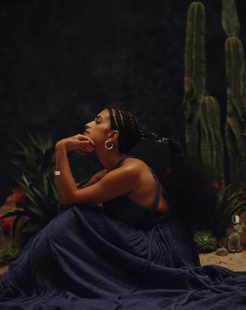 Elegant woman in flowing blue dress poses thoughtfully in a desert-like setting with cacti and dark background, illuminated by soft light.