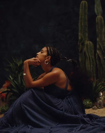 Elegant woman in flowing blue dress poses thoughtfully in a desert-like setting with cacti and dark background, illuminated by soft light.