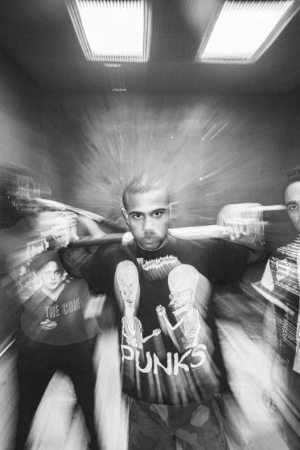 Black and white shot of an adult male wearing a t-shirt with a graphic print standing indoors with two people on either side.