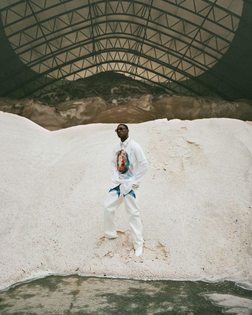 A stylish Black man in white clothing stands in a quarry-like environment, under a large industrial dome, surrounded by white material.