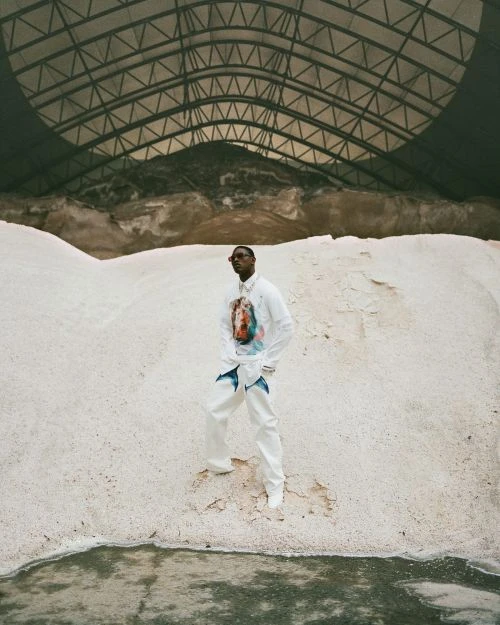 A stylish Black man in white clothing stands in a quarry-like environment, under a large industrial dome, surrounded by white material.