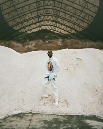 A stylish Black man in white clothing stands in a quarry-like environment, under a large industrial dome, surrounded by white material.
