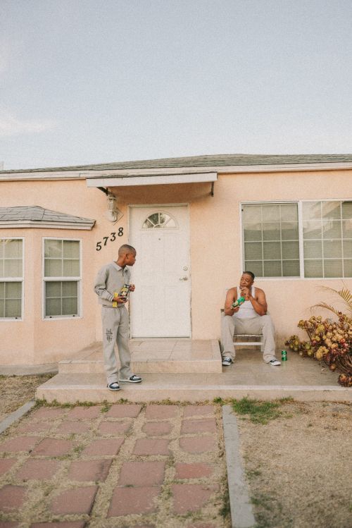 Two men in casual clothing sit on the porch of a peach-colored house on a sunny day, one holding a beverage and a snack.