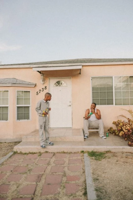 Two men in casual clothing sit on the porch of a peach-colored house on a sunny day, one holding a beverage and a snack.