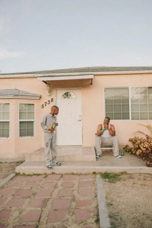 Two men in casual clothing sit on the porch of a peach-colored house on a sunny day, one holding a beverage and a snack.