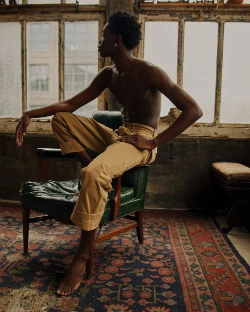 Adult Black man with bare chest sits on a green chair in front of a window wearing tan pants and silver earrings in an indoor setting.