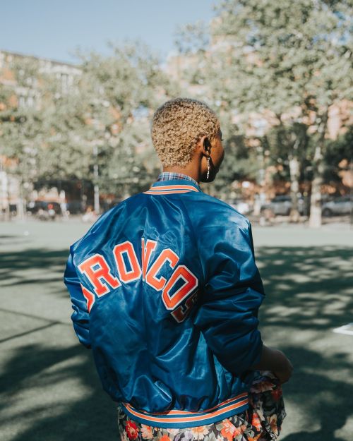 Adult woman with blond hair in blue and orange 'Broncos' satin jacket outside on a sunny day with trees and buildings in the background.
