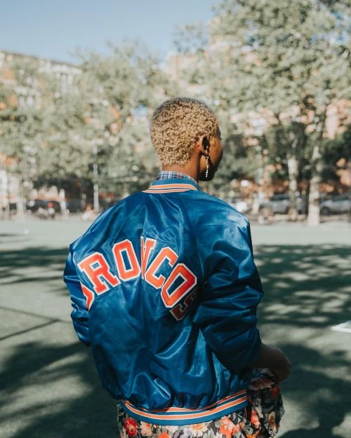 Adult woman with blond hair in blue and orange 'Broncos' satin jacket outside on a sunny day with trees and buildings in the background.