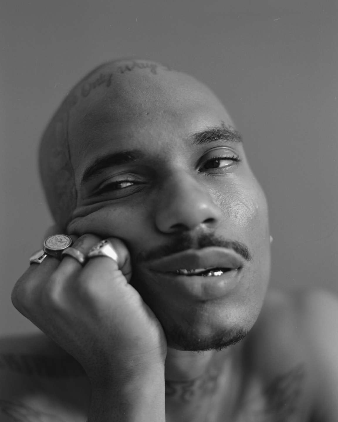 Monochrome portrait of a bald adult male with a mustache, tattoos, and rings posing indoors, resting his face on his hand.