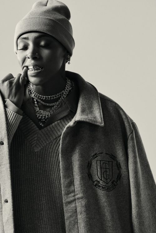 Portrait of a young adult woman in a beanie and grey outerwear, with dental braces and layered necklaces against a neutral backdrop.