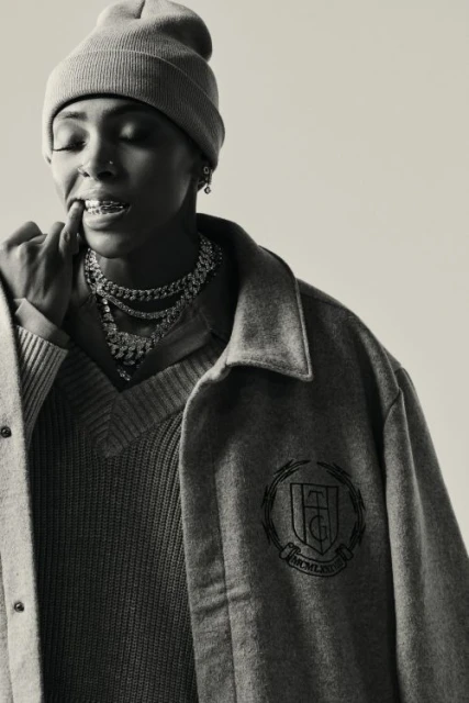 Portrait of a young adult woman in a beanie and grey outerwear, with dental braces and layered necklaces against a neutral backdrop.