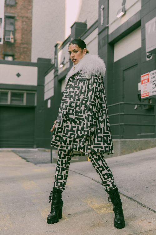 Adult female fashion model poses in a patterned coat and matching leggings in an urban outdoor setting with an expressive facial expression.