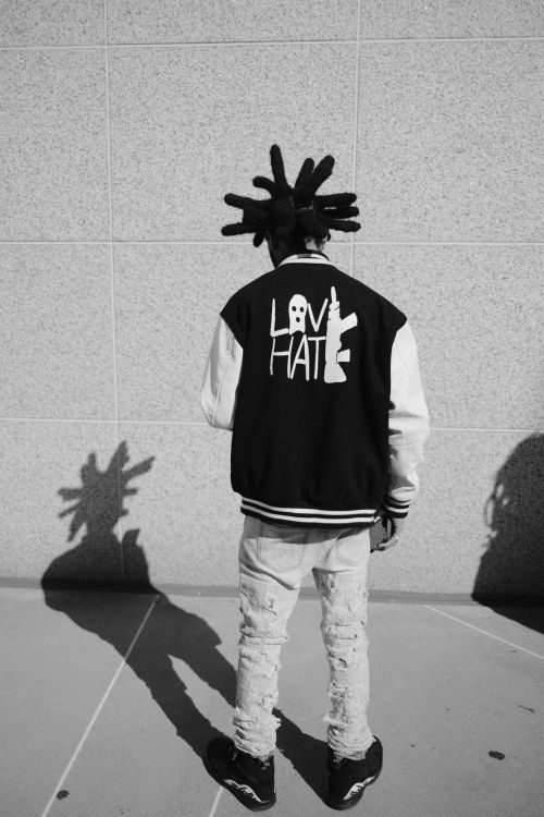 Black-and-white shot of a person with dreadlocks wearing a jacket with 'LOVE' and 'HATE' graphics, standing in front of a textured wall.