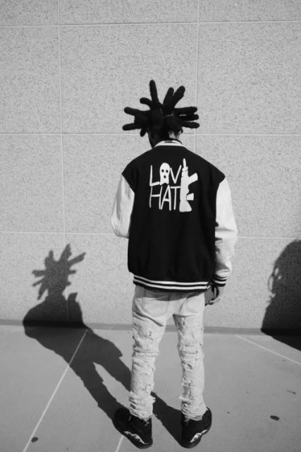 Black-and-white shot of a person with dreadlocks wearing a jacket with 'LOVE' and 'HATE' graphics, standing in front of a textured wall.