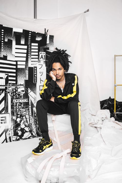 A young Black man with locs seated indoors wearing a black tracksuit with yellow stripes and matching sneakers in front of a backdrop.