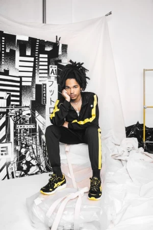 A young Black man with locs seated indoors wearing a black tracksuit with yellow stripes and matching sneakers in front of a backdrop.
