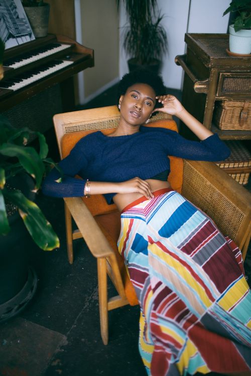 A young adult woman with dark hair is reclining in an armchair with colorful skirt; potted plant, cabinet, and piano in background.