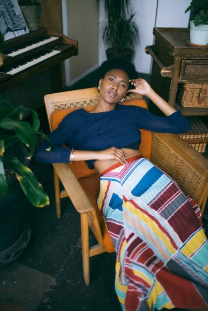 A young adult woman with dark hair is reclining in an armchair with colorful skirt; potted plant, cabinet, and piano in background.