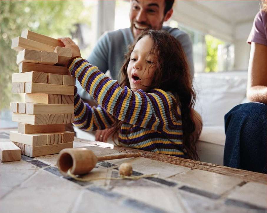 Child carefully removes block from wooden tower game, watched by smiling father and seated adult, in a bright indoor setting.