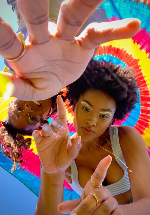 Two African American women pose in an upward shot; one displays makeup with a colorful tie-dye background and gold rings on fingers.