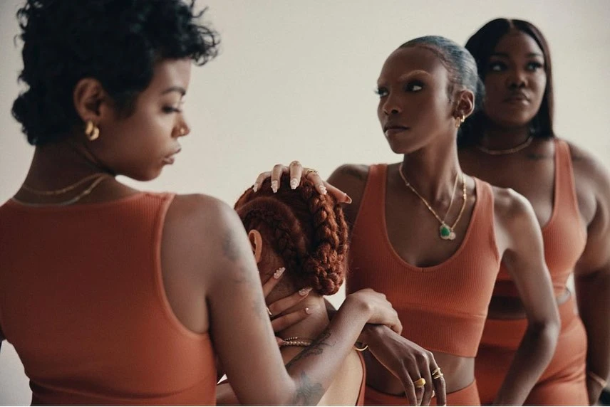 Four African American women pose in matching outfits in a studio, displaying fashionable gold jewelry against a plain backdrop.