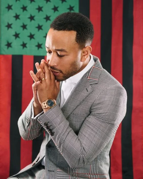 Portrait of John Legend in grey suit and white shirt, hands clasped near his face, set against an Afro-American flag backdrop.