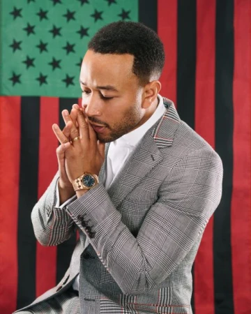 Portrait of John Legend in grey suit and white shirt, hands clasped near his face, set against an Afro-American flag backdrop.