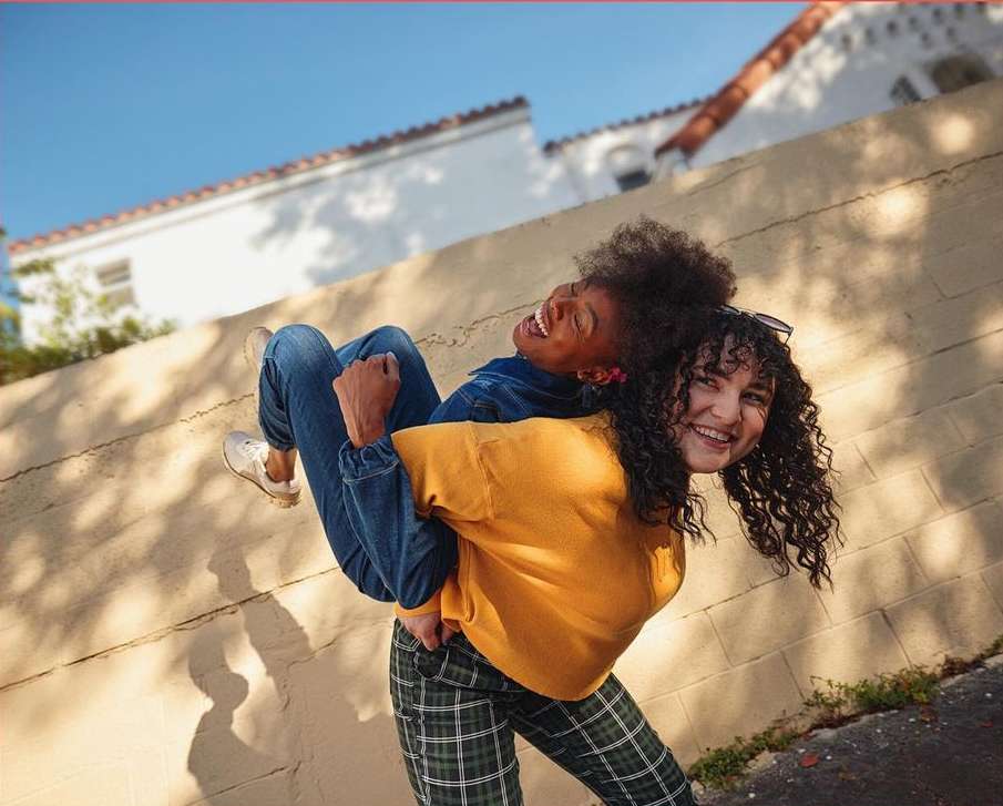 Two young women laugh and smile in sunlight, one giving the other a piggyback ride, with blue sky and buildings in the background.