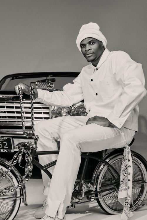An adult man poses on a vintage bicycle, wearing a white outfit and matching hat, with a vintage car partially visible in the background.