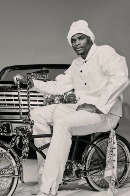 An adult man poses on a vintage bicycle, wearing a white outfit and matching hat, with a vintage car partially visible in the background.