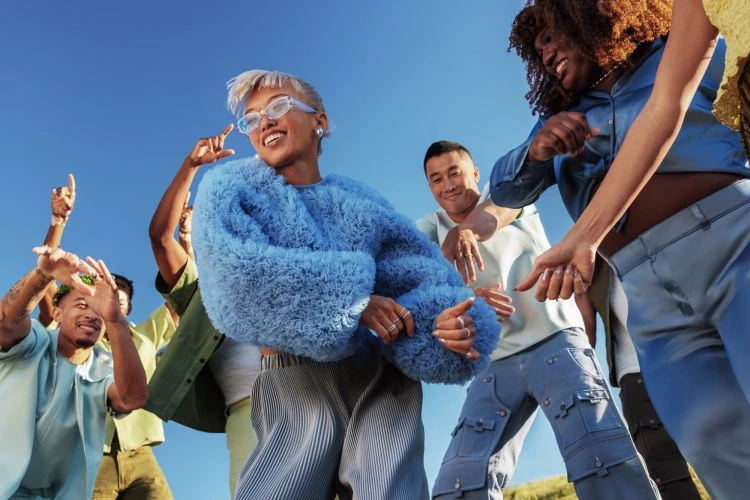 A group of diverse young adults dance and enjoy a bright, sunny day. They wear stylish clothing in a casual outdoor setting.