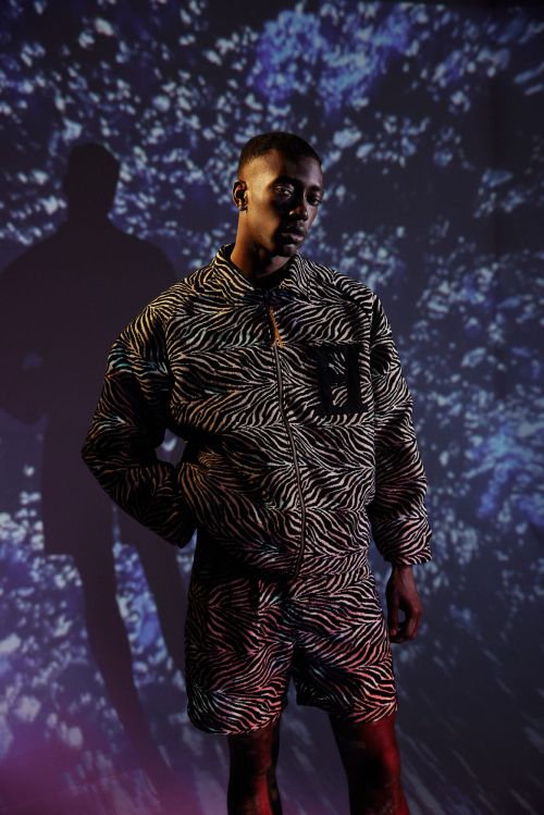 Adult male in zebra-striped jacket and shorts against a blue floral background with shadows. He is looking off camera with hand in pocket.