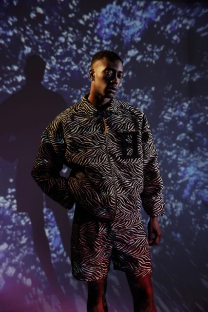 Adult male in zebra-striped jacket and shorts against a blue floral background with shadows. He is looking off camera with hand in pocket.