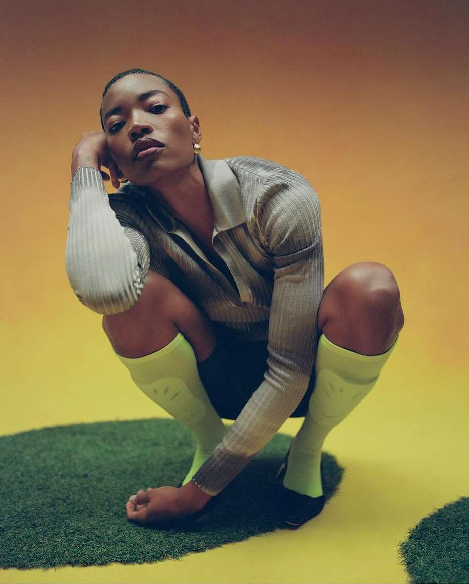 Woman in grey ribbed shirt and yellow socks crouches on a patch of grass with a yellow and orange background in a studio setting.