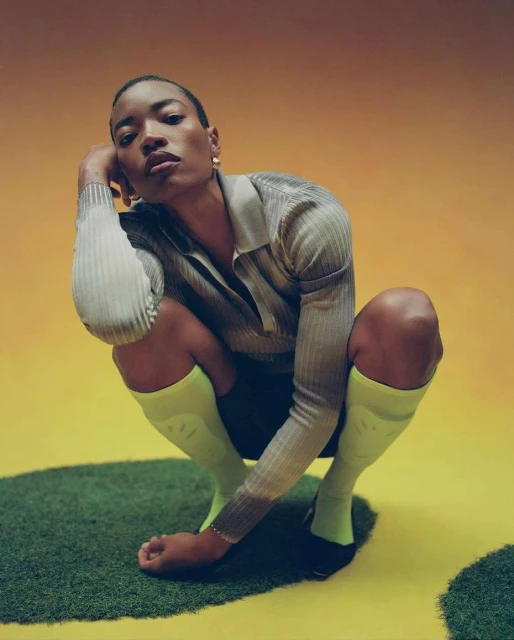 Woman in grey ribbed shirt and yellow socks crouches on a patch of grass with a yellow and orange background in a studio setting.