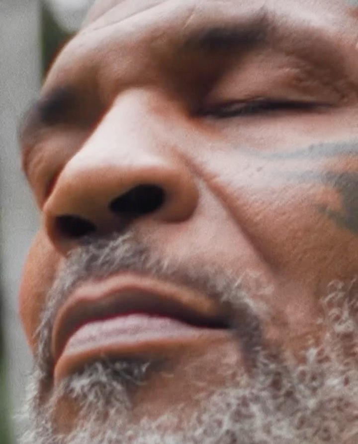 Close-up shot of an adult male's face with a partially closed eye, a gray beard, and face tattoo against a blurred, natural background.