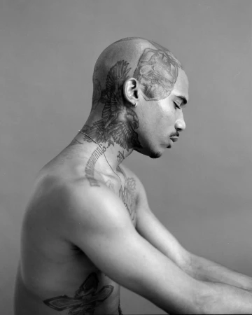 Profile portrait of a shirtless man with extensive head, neck, and torso tattoos against a solid grey background