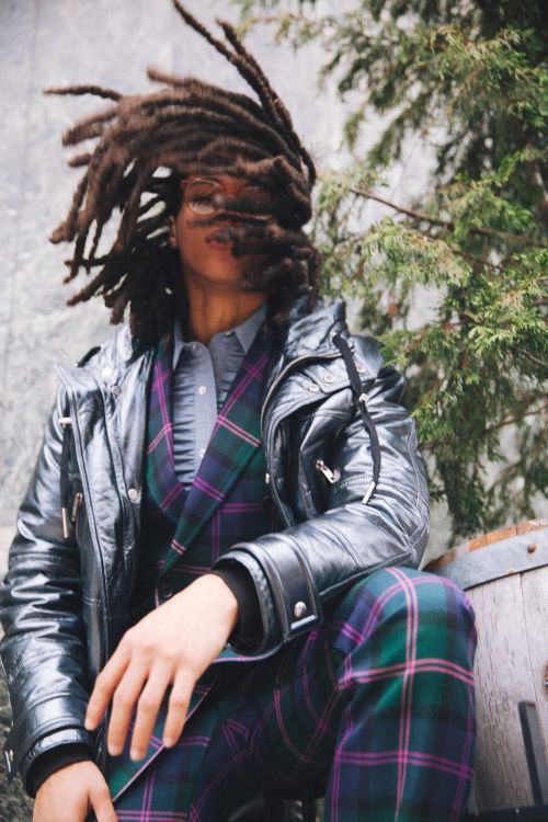 Portrait of a person with dreadlocks wearing a green plaid suit and leather jacket sitting outside near evergreen branches and a stone wall.