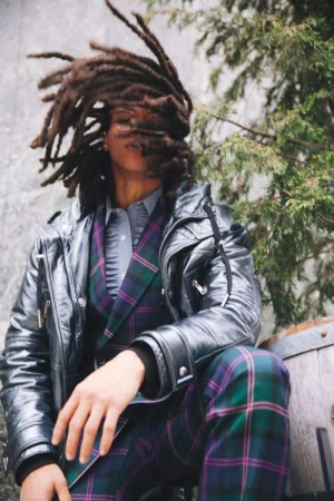 Portrait of a person with dreadlocks wearing a green plaid suit and leather jacket sitting outside near evergreen branches and a stone wall.