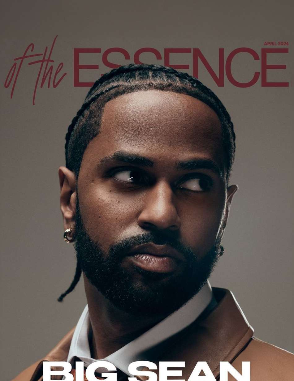 Portrait of rapper Big Sean with braided hair and a beard, wearing a brown jacket over a white collared shirt against a grey background.