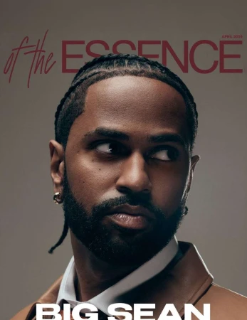 Portrait of rapper Big Sean with braided hair and a beard, wearing a brown jacket over a white collared shirt against a grey background.