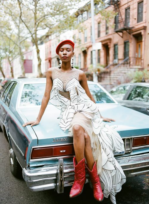 Fashionable woman in red beret and boots poses on classic light blue car in urban setting with brownstones in background.