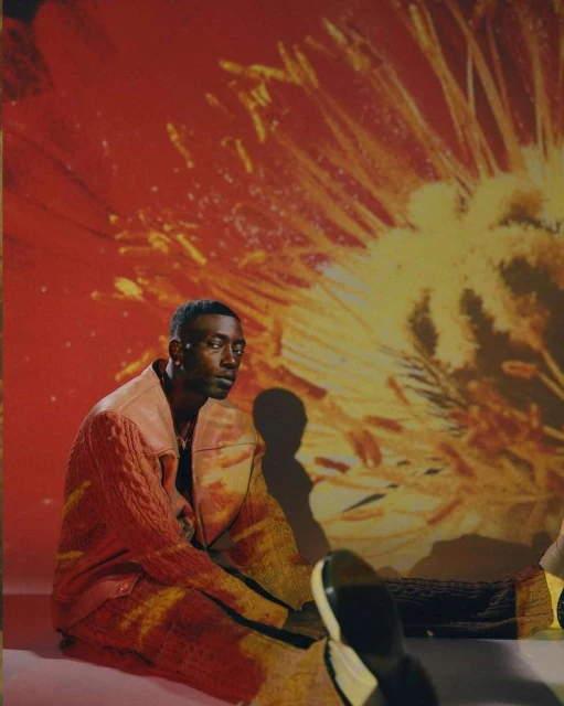 A stylish African-American man poses in a pink and orange knit jacket, seated against an abstract floral background with fashion-forward appeal.